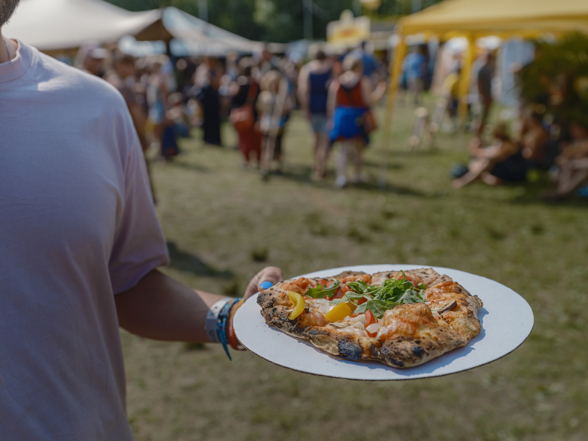 Close-up of a person in a white t-shirt holding a paper plate with a freshly-made artisanal pizza on an outdoor food festival