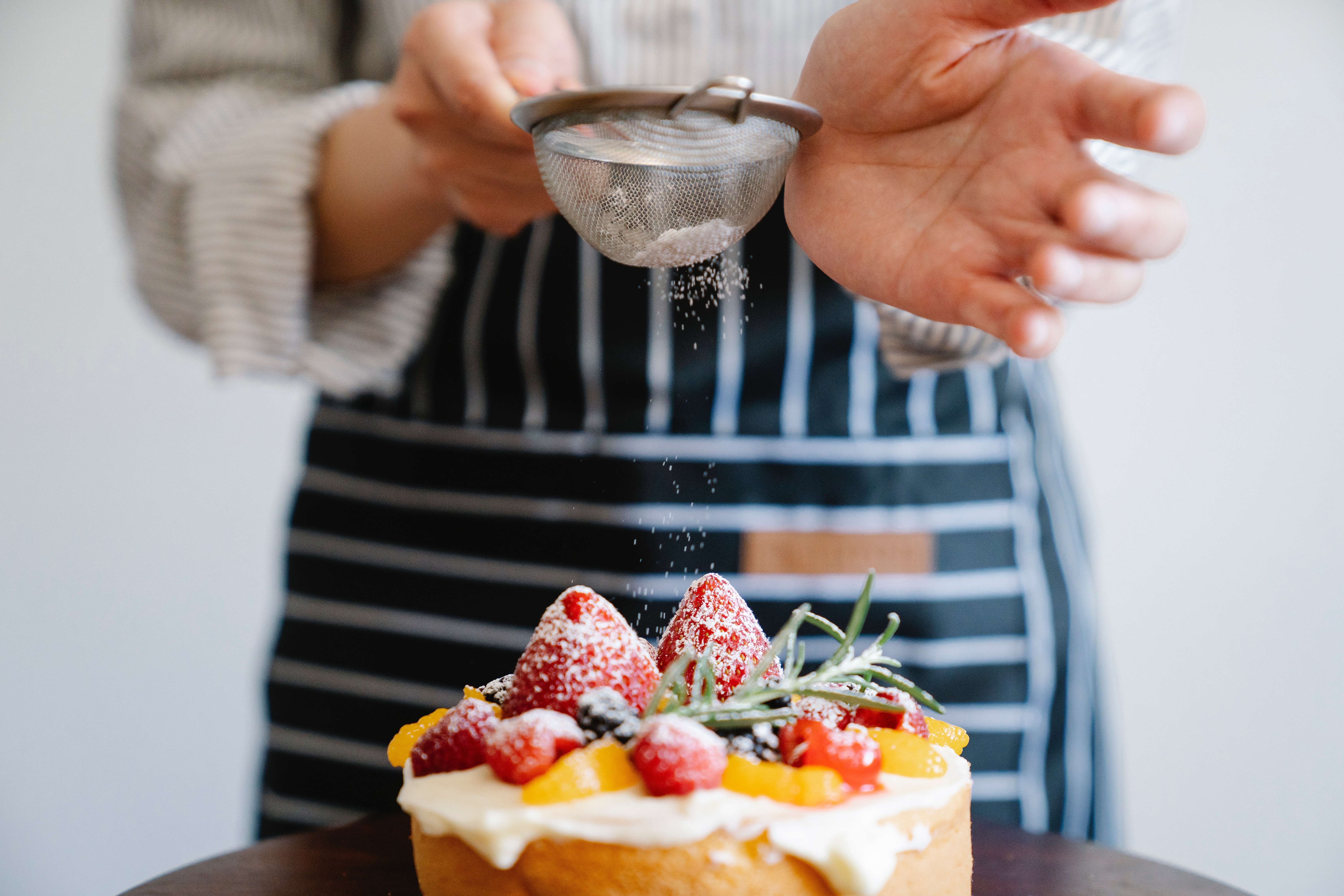 man in a striped apron sprinkling powdered sugar over a cake with strawberries